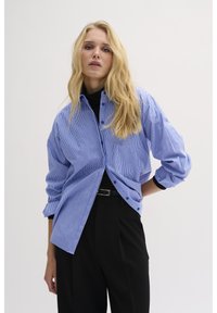 Blue and white striped button-up shirt with a relaxed fit, long sleeves, and contrasting black accents. Paired with black trousers.