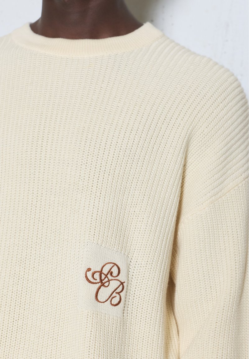 Pull tricoté couleur crème avec une texture côtelée et un petit patch brodé avec des lettres cursives "S" et "B" sur la poitrine.