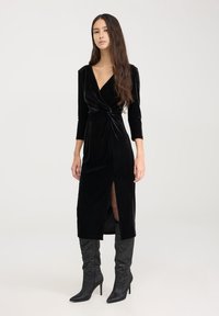 Czarna aksamitna sukienka z długimi rękawami, detalem w formie skrętu w talii oraz bocznym rozcięciem. Połączona z błyszczącymi czarnymi kozakami do kolan.