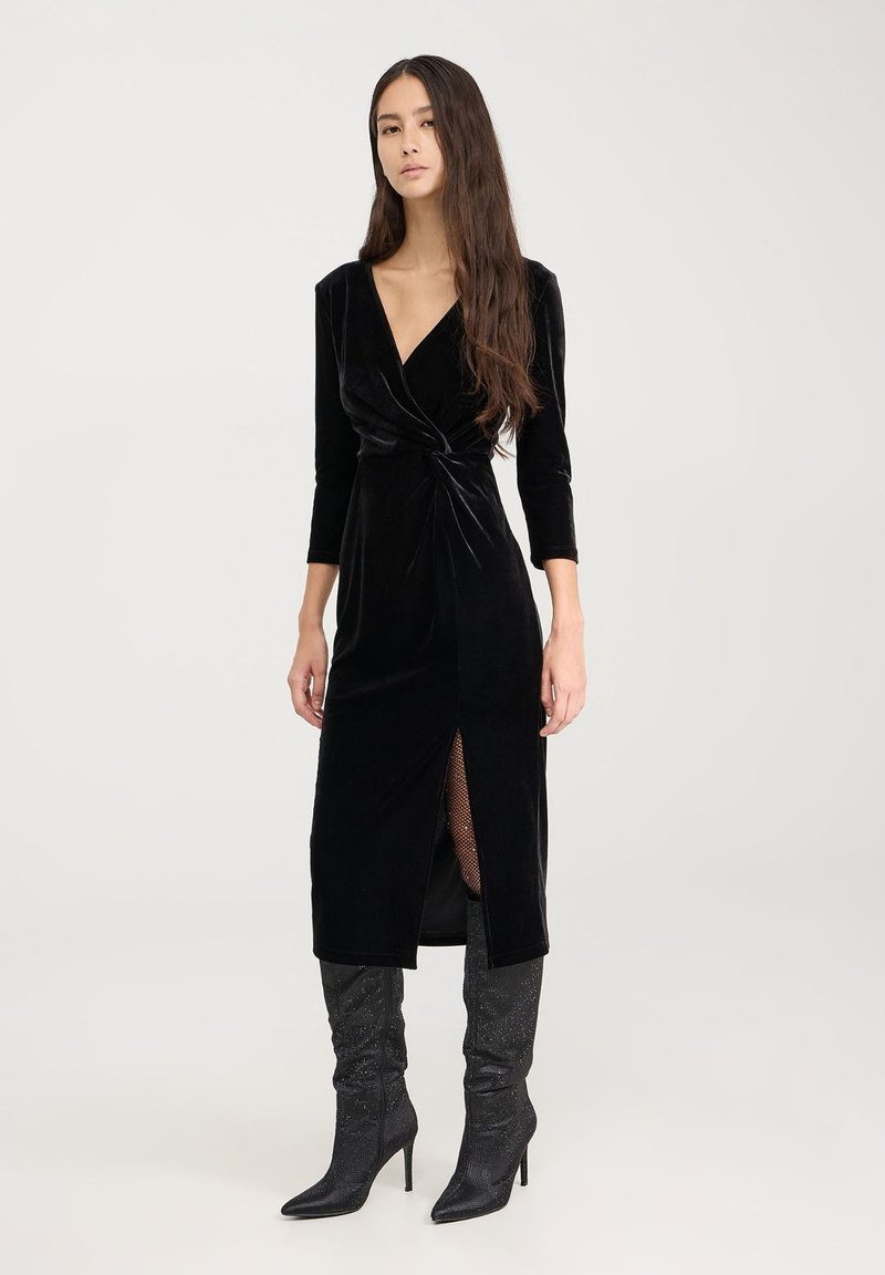 Czarna aksamitna sukienka z długimi rękawami, detalem w formie skrętu w talii oraz bocznym rozcięciem. Połączona z błyszczącymi czarnymi kozakami do kolan.