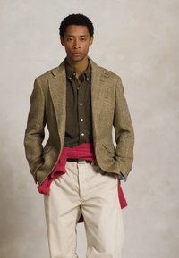 Blazer de tweed castanho, camisa escura, calças cremes e um suéter rosa atado à cintura, apresentando um corte clássico e texturas.