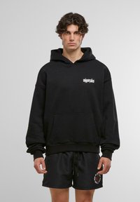 Hanorac negru cu glugă, având o buzunar frontal, cu text alb pe piept. Purtați cu pantaloni scurți negri care prezintă o broderie decorativă.