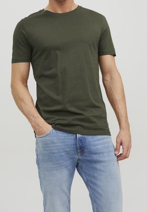 Mand iført olivengrøn kortærmet T-shirt og lyseblå jeans, står med den ene hånd i lommen mod en ensfarvet baggrund.