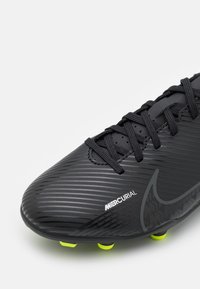 Botas de fútbol Nike Mercurial negras con superficie texturizada, cordones negros y tacos amarillos neón sobre un fondo blanco.