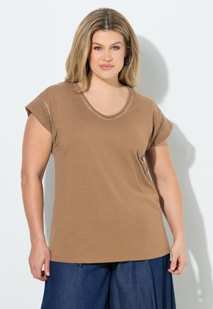 Woman with shoulder-length blonde hair wearing a light brown short-sleeve top and dark blue wide-leg pants, standing against a white wall.