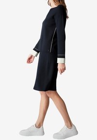 Navy-blaues Strickkleid mit gerader Silhouette, weißen Ärmelakzenten und Seitenpipings. Kombiniert mit weißen Sneakers. Glatte Textur.