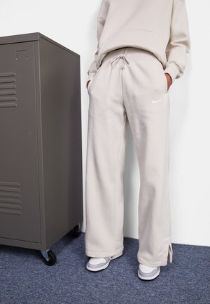 Personne portant un pantalon de survêtement beige Nike et un sweat assorti, debout à côté d'un casier métallique gris sur un sol moquetté bleu.