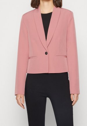 Kobieta nosząca dopasowaną marynarkę w kolorze pudrowego różu z jednym guzikiem, na czarnym topie i czarnych spodniach, pokazana od szyi do bioder na białym tle.