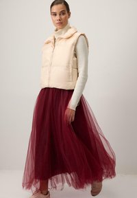 Pūkains krēmkrāsas veste ar priekšējo rāvējslēdzēju pār gariem bordo tilla svārkiem. Apģērbā ir piegulošs augsta kakla džemperis zem tā.