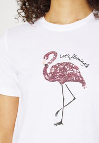 Biała koszulka z różowym, cekinowym nadrukiem flaminga. Tekst "Let's flamingle" w czarnym napisie, z prostymi rysunkami jako szczegóły.