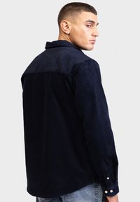 Chemise en velours côtelé bleu marine avec un col, des poignets à boutons et une texture côtelée. Présente un design de piece distinctive au dos.