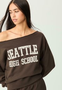 Felpa marrone oversize con design off-shoulder, caratterizzata dalla scritta "SEATTLE HIGH SCHOOL" in bianco sul davanti. Texture morbida.