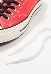 Baskets en toile rouge avec lacets noirs et œillets en argent, dotées d'une coque de protection en caoutchouc blanc et d'une semelle texturée blanche. Comprend un lacet blanc.