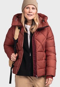Rote gesteppte Daunenjacke mit abnehmbarer Kapuze, Reißverschluss vorne und Seitentaschen. Getragen über einer schwarzen Fleece-Schicht, ergänzt durch einen beigen Rucksack.