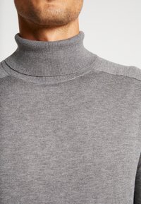 Grå turtlenecktröja i mjukt stickat material. Har en ribbad halsringning och en slät yta, med minimal detaljering på axlarna.