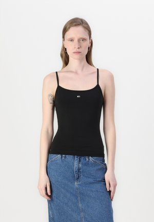 Γυναίκα με ανοιχτόχρωμο δέρμα και ίσια καστανά μαλλιά φοράει μαύρο τοπ με λεπτές τιράντες και μπλε τζιν φούστα, στέκεται μπροστά σε ένα απλό λευκό φόντο.