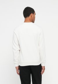 Hombre de espaldas vistiendo un sudadera blanca de manga larga y pantalones negros frente a un fondo blanco.