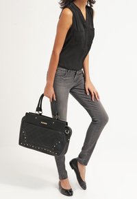 Femme portant un haut noir sans manches, un jean skinny gris, des ballerines noires, tenant un grand sac à main matelassé noir avec des clous dorés.