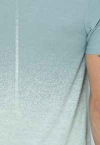 Lyseblå gradient t-shirt med en tekstureret finish, der går fra ensfarvet til et plettet mønster nær kanten. Korte ærmer.