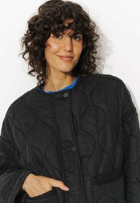 Veste matelassée noire avec un motif texturé, des boutons-pression et des poches latérales. Portée sur un haut bleu, montrant une coupe décontractée.