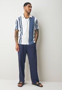 Kurzärmeliges, weißes und marineblau gestreiftes Hemd mit Reißverschluss, kombiniert mit locker sitzenden, marineblauen Hosen und schwarzen Sandalen.