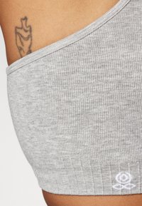 Gros plan sur l'épaule d'une personne portant un haut gris côtelé à une épaule avec un petit logo blanc brodé près de l'ourlet et un tatouage visible.