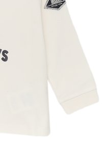T-shirt a maniche lunghe colore crema con scollo rotondo, caratterizzata da una grafica testuale blu sul davanti e una macchia scura con testo bianco sulla manica.