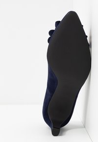 Chaussure en daim bleu marine à bout pointu avec un talon fin, vue de la semelle noire sur fond blanc.