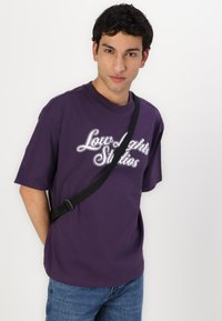Ljubičasta pamučna majica s bijelom vezom na kojoj piše "Low Lights Studios", uparen s svijetloplavim trapericama i crnim crossbody remenom.