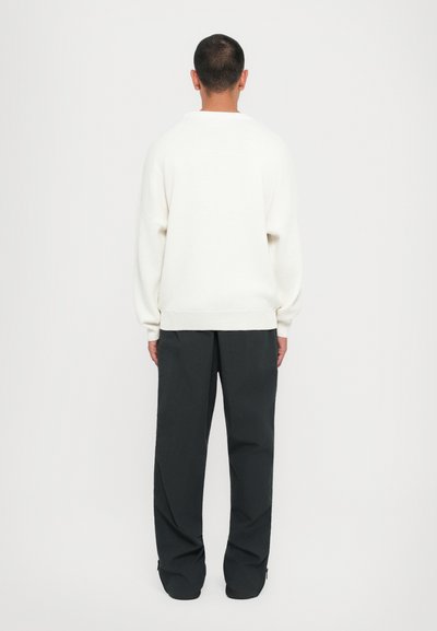 Weißer Strickpullover mit lockerer Passform, überschnittenen Schultern und gerippten Bündchen. Kombiniert mit schwarzen weiten Hosen, die eine glatte Textur aufweisen.