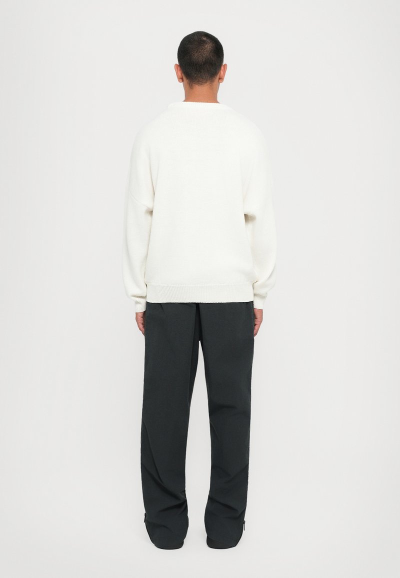 Pull en tricot blanc à coupe décontractée, avec des épaules tombantes et des poignets côtelés. Associé à un pantalon large noir, doté d'une texture lisse.