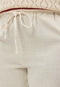 Nærbilde av lyse beige bukser med en tekstil snor knyttet i midjen og en teksturert kremhvit genser som er stukket inn.