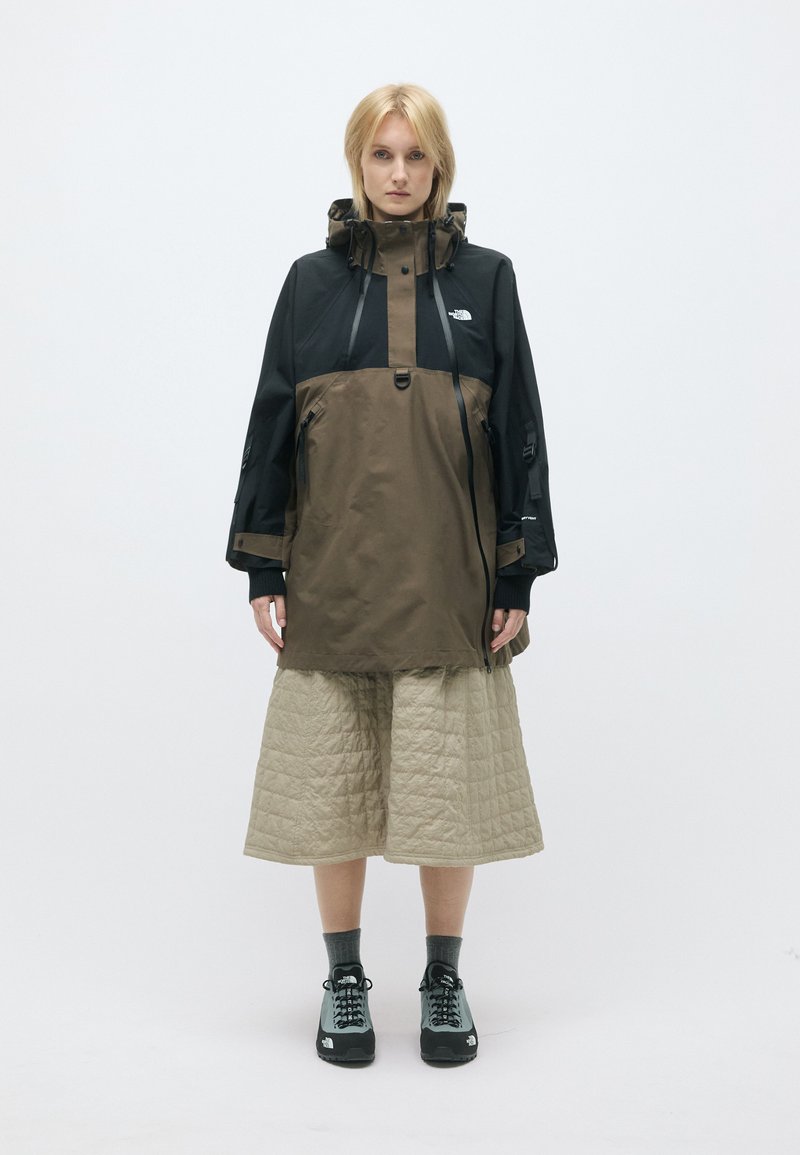 Brūna un melna ūdensizturīga jaka ar augstu apkakli, priekšējiem kabatām un regulējamām siksnām, kombinēta ar polsterētu beigu svārku un melniem apaviem.