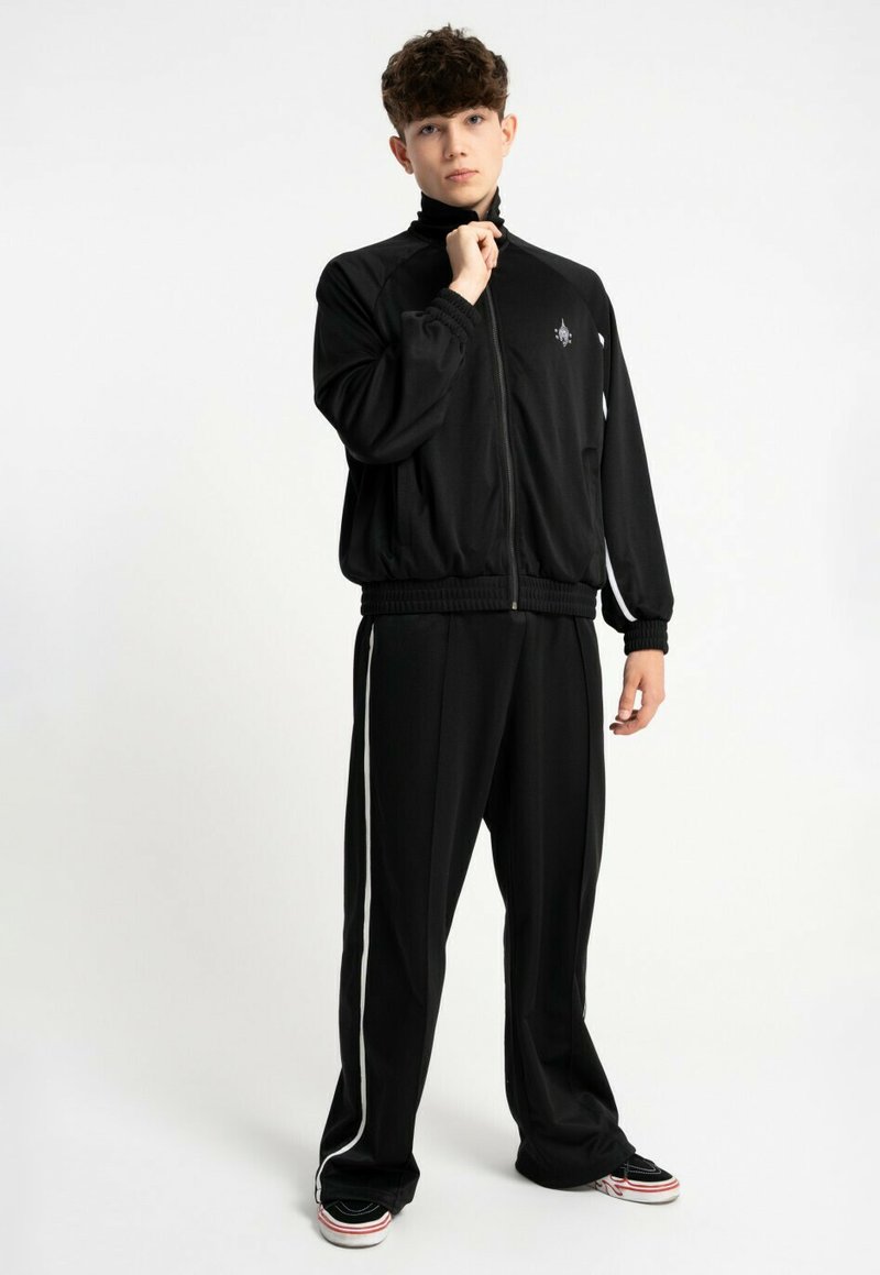 Młoda osoba stojąca w czarnym dresie z białymi paskami, w zapinanej kurtce, czarnych sneakersach z białymi podeszwami na jednolitym białym tle.