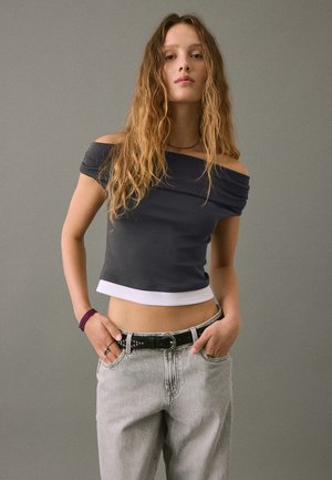 Mlada ženska z dolgimi valovitimi lasmi, oblečena v odprto črno crop top majico, svetlo sivo jeans hlače, črn pas in vijoličen beaded zapestnico, stoji z rokama v žepih.