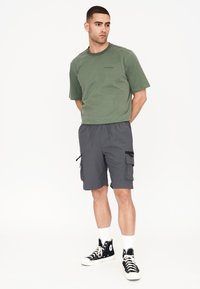 Grön bomull t-shirt, kort ärm, rundad halsringning; grå cargo shorts med dragkedjefickor; svarta hög-top sneakers med vita snören.