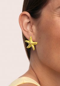 Pendiente en forma de estrella de mar de color amarillo con acentos en tono dorado, que presenta una textura suave y cinco puntas salientes, llevado en la oreja.