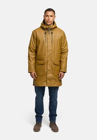Impermeabile giallo-marrone con cappuccio, dotato di due tasche frontali, chiusura con coulisse e una texture liscia e lucida. Indossato sopra a dei jeans.