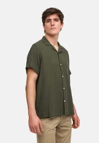 Chemise à manches courtes, de couleur olive, à boutons, fabriquée en tissu texturé, avec un col cranté et cinq boutons blancs à l'avant.