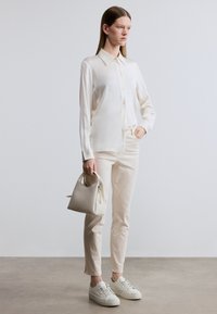 Κρεμ μεταξωτό πουκάμισο με κουμπιά, μπεζ εφαρμοστά παντελόνια και λευκά αθλητικά παπούτσια. Κρατάει μια μικρή κρεμ τσάντα χειρός με φερμουάρ. Λιτές υφές, μίνιμαλ σχεδιασμός.