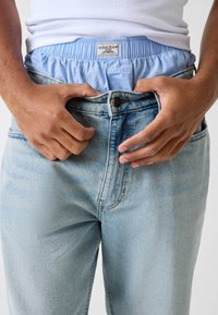 Jean en denim bleu clair avec une coupe décontractée, doté d'un design à cinq poches et d'une fermeture à boutons. Taille élastique visible et étiquette.