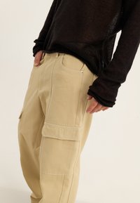 Beige cargo hlače s sproščenim krojem, ki imajo stranske žepe in bele šive. Ujemajo se z ohlapno, črno pleteno majico z dolgimi rokavi.
