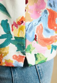 Detailný záber na látku s kvetinovým vzorom s viacfarebnými kvetmi v ružovej, oranžovej, modrej a zelenej na bielej, položenú na modrých rifliach.