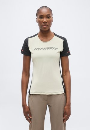 Γυναίκα που φοράει κοντομάνικη αθλητική μπλούζα Dynafit σε μπεζ και μαύρο χρώμα και ανοιχτό καφέ παντελόνι, στέκεται μπροστά σε απλό φόντο.