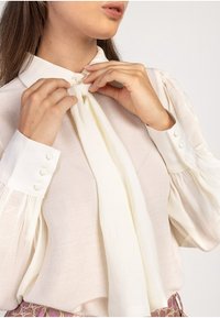 Vit blus med hög krage och knytdetalj, med långa ärmar med knappdekorationer. Strukturerad tyg med en subtil lyster.