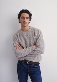Maglione grigio chiaro a maglia con texture a coste, abbinato a jeans in denim blu scuro e una cintura marrone. Il modello è in piedi con le braccia incrociate.
