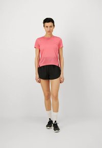 Růžové tričko s krátkým rukávem pro sport, z lehké látky, se štěrbinami po stranách, v kombinaci s černými kraťasy, bílými ponožkami a černými teniskami.