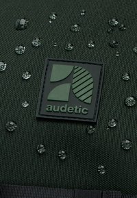 Zelená látka s texturovaným povrchem obsahuje kapky vody a černou nášivku s nápisem "audetic" s geometrickými vzory.