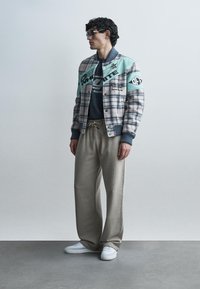 Homme portant une veste universitaire à motifs, un t-shirt sombre, un pantalon beige à cordon, des baskets blanches et des lunettes de soleil noires, debout devant un fond uni.