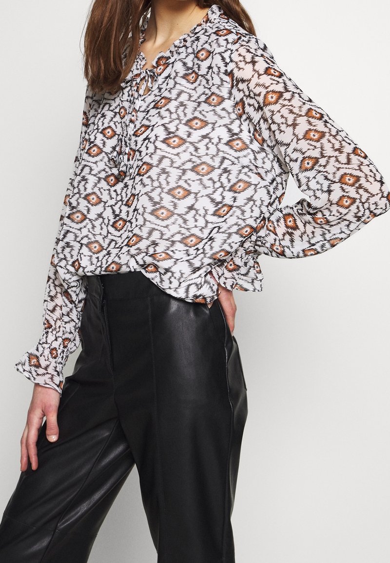 Transparante witte blouse met een abstract oranje en zwart geometrisch patroon, lange mouwen met elastische manchetten, en een strikdetail bij de halslijn.